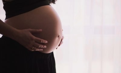 Eine schwangere Frau steht vor einem Fenster. Sie hält sich mit beiden Händen den Bauch.