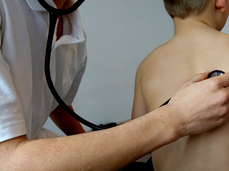 Eine Hand eines Mannes (Kinderarzt) mit weißem T-Shirt hält ein Stethoskop an den Rücken eines Kindes.p