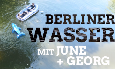 Ein aufblasbares Boot ist auf der Spree. Darin sitzen die Abgeordneten June Tomiak und Georg Kössler. Daneben schwimmt ein blauer aufblasbarer Delfin. Auf der Wasserfläche daneben ist der Text "Berliner Wasser mit June und Georg" zu lesen.