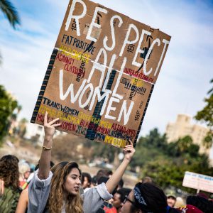 Es ist eine Frau auf einer Demonstration zu sehen, die ein Schild mit der Aufschrift "Respect all women" hoch hält.