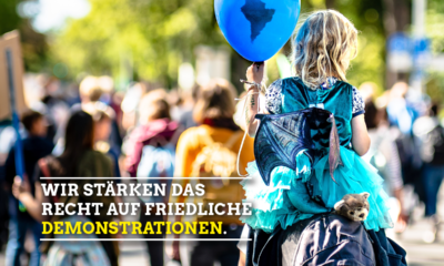 Bild von einer Demonstration von den Rücken der Menschen. Im Vordergrund sitzt ein blondes Mädchen auf den Schultern eines Elternteils und hält einen blauen Luftballon in der Hand. In der linken Hand steht in weißen Buchstaben wir stärken das Recht auf friedliche und in gelb Demonstrationen.