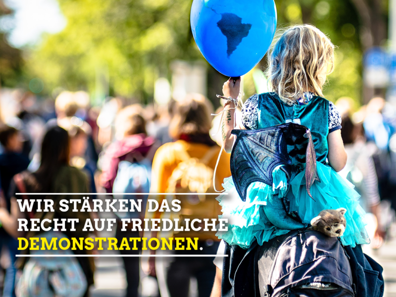 Bild von einer Demonstration von den Rücken der Menschen. Im Vordergrund sitzt ein blondes Mädchen auf den Schultern eines Elternteils und hält einen blauen Luftballon in der Hand. In der linken Hand steht in weißen Buchstaben wir stärken das Recht auf friedliche und in gelb Demonstrationen.