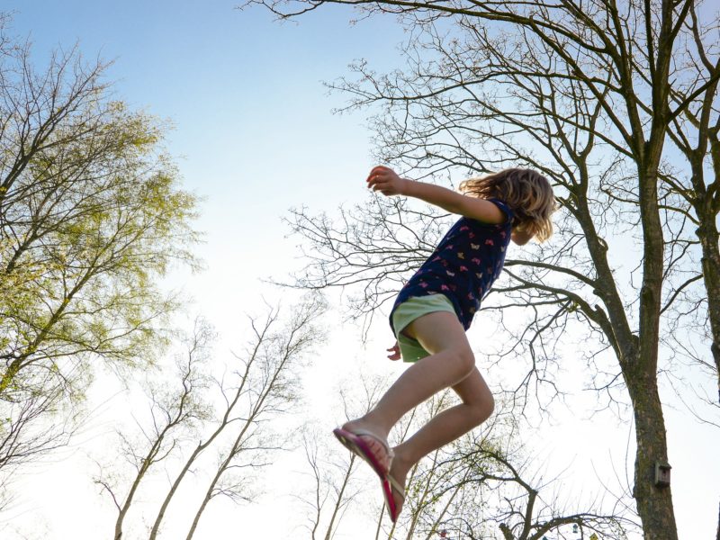 Kind springt auf einem Trampolin in die Höhe; das Foto ist so gemacht worden, dass nur ein Kind und Bäume, die in den Himmel ragen, zu sehen sind.