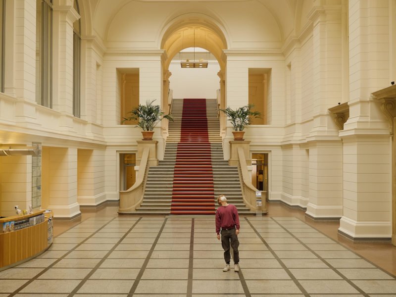 Auf dem Bild ist die Eingangshalle des Abgeordnetenhauses zu sehen in deren Mitte eine Person mit brauner Hose und rotem Pulli steht, die in die Höhe schaut.