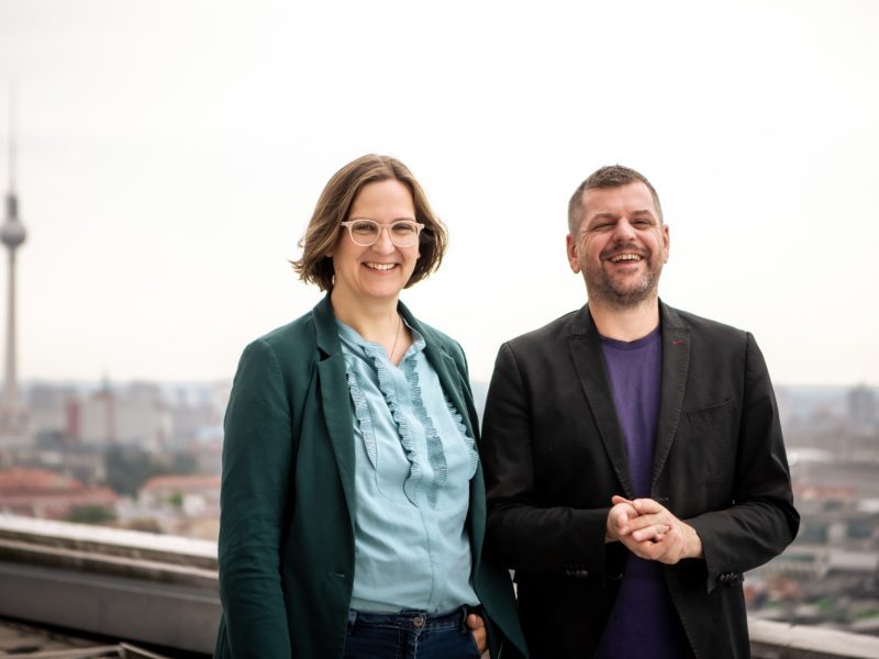 Porträt von Silke Gebel und Werner Graf. Sie sind seriös angezogen und stehen auf einem Berliner Dach, im Hintergrund der Fernsehturm. Beide lächeln in die Kamera.