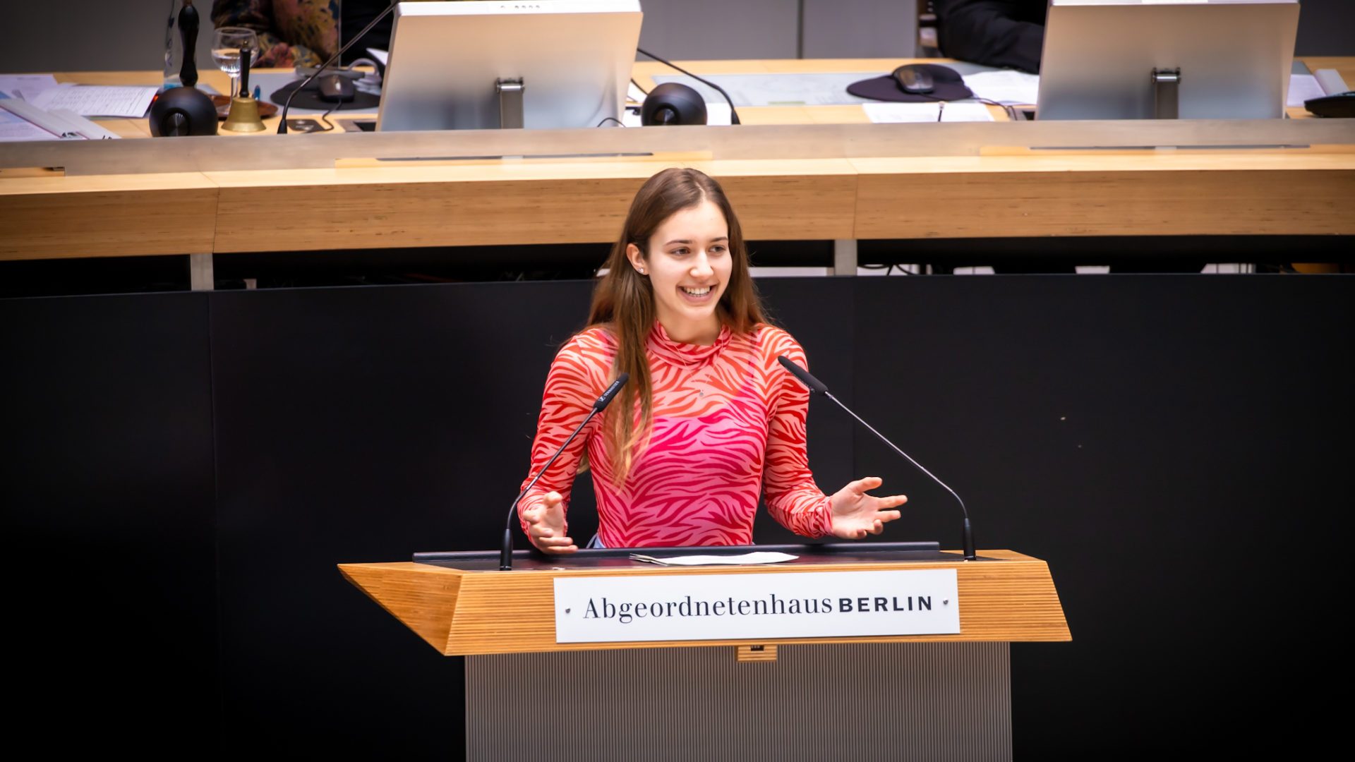 Klara Schedlich steht am Redepult des Abgeordnetenhauses. Sie lächelt und breitet beide Hände aus. Sie ist sehr jung und trägt ein pinkes Oberteil.