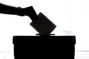 Foto von einer Wahlurne, in die ein Zettel gesteckt wird. Sie liegt komplett im Schatten und ist daher schwarz.
