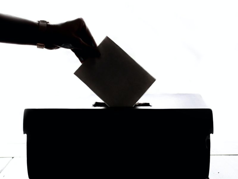 Foto von einer Wahlurne, in die ein Zettel gesteckt wird. Sie liegt komplett im Schatten und ist daher schwarz.
