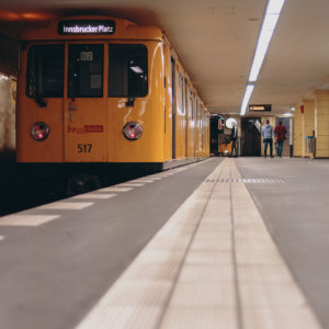 Von unten aufgenommen zeigt das Bild einen U-Bahnhof von innen. Links ist eine orangene U-Bahn zusehen, rechts ist der Bahnsteig zu sehen. Er hat einen grauen Boden und eine weiße Linie als Leitsystem.