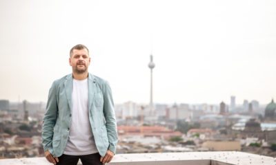 Werner Graf steht auf den Dächern Berlins, im Hintergrund sieht man den Fernsehturm. Er trägt ein hellblaues Jackett und steht sehr stabil da. Er schaut entschlossen.