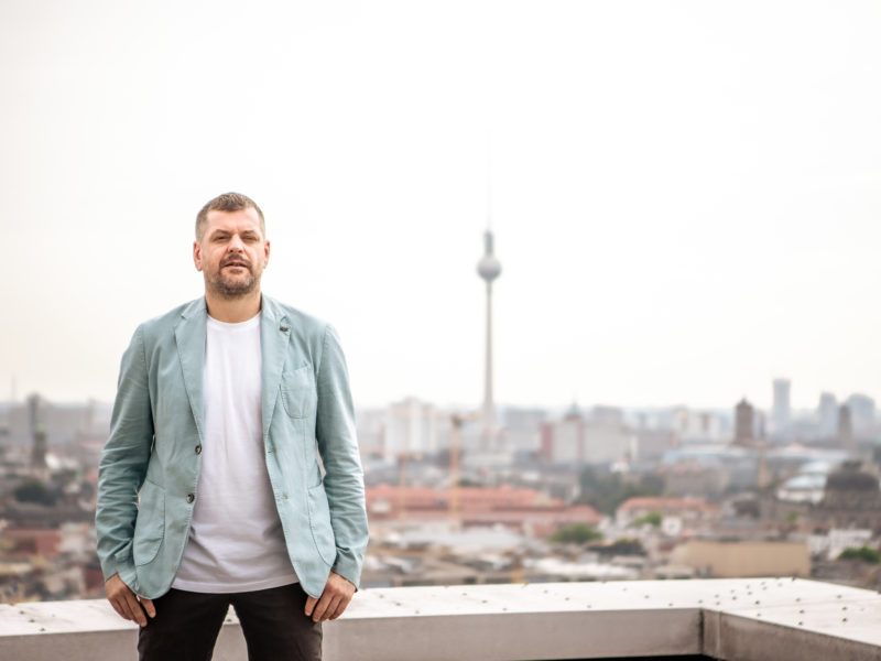 Werner Graf steht auf den Dächern Berlins, im Hintergrund sieht man den Fernsehturm. Er trägt ein hellblaues Jackett und steht sehr stabil da. Er schaut entschlossen.