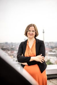 Silke Gebel steht auf einem Dach Berlins, die Arme verschärnkt. Sie trägt einen orangefarbenen Jumpsuit und einen schwarzen Blazer. Sie lächelt in die Kamera. Im Hintergrund sieht man den Fernsehturm.