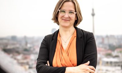 Silke Gebel steht auf einem Dach Berlins, die Arme verschärnkt. Sie trägt einen orangefarbenen Jumpsuit und einen schwarzen Blazer. Sie lächelt in die Kamera. Im Hintergrund sieht man den Fernsehturm.