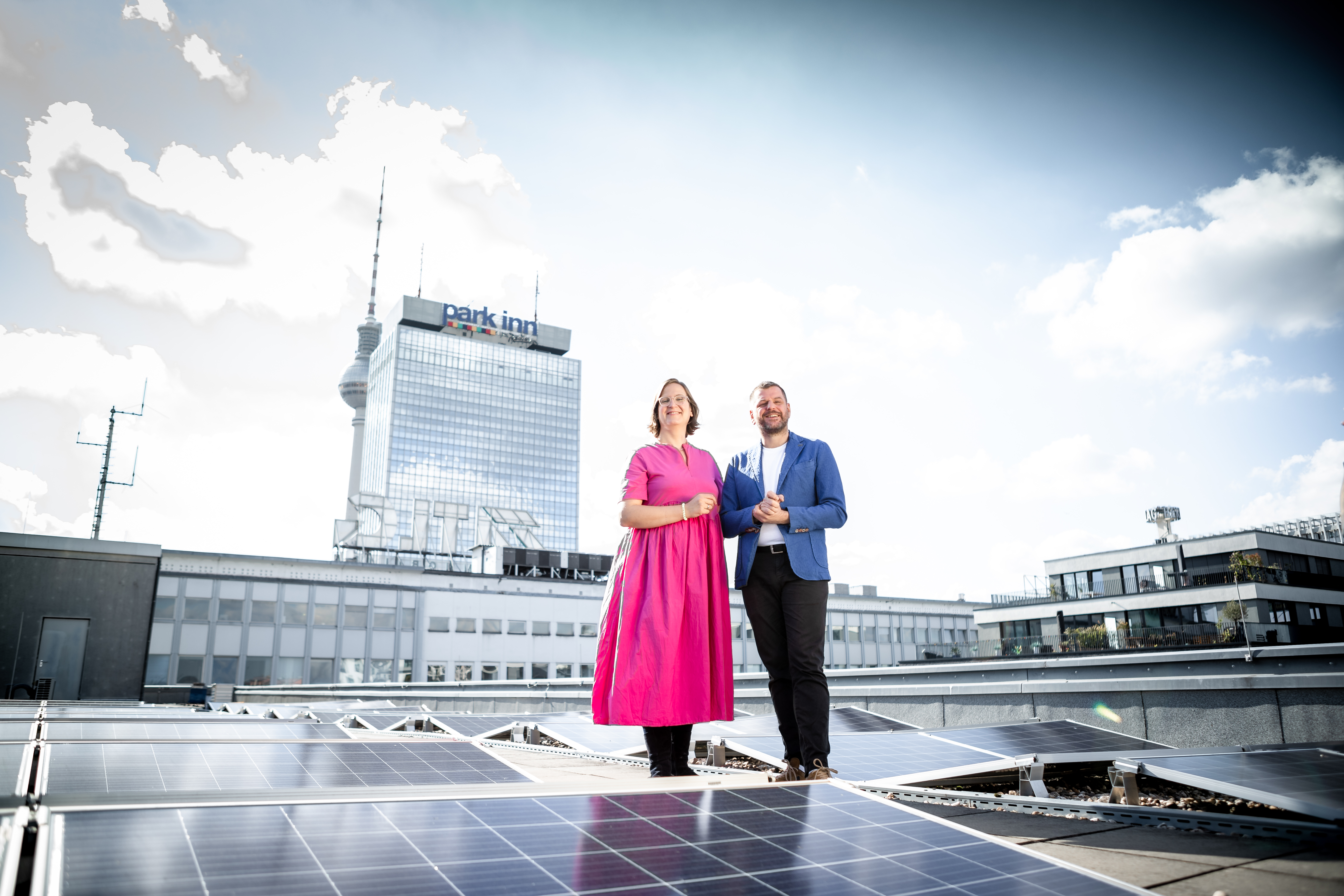 Silke Gebel und Werner Graf stehen auf einem Flachdach, um sie herum sind Solarpanels. Silke trägt ein leuchtend pinkes Kleid. Im Hintergrund sieht man ein Hochhaus und den Fernsehturm.