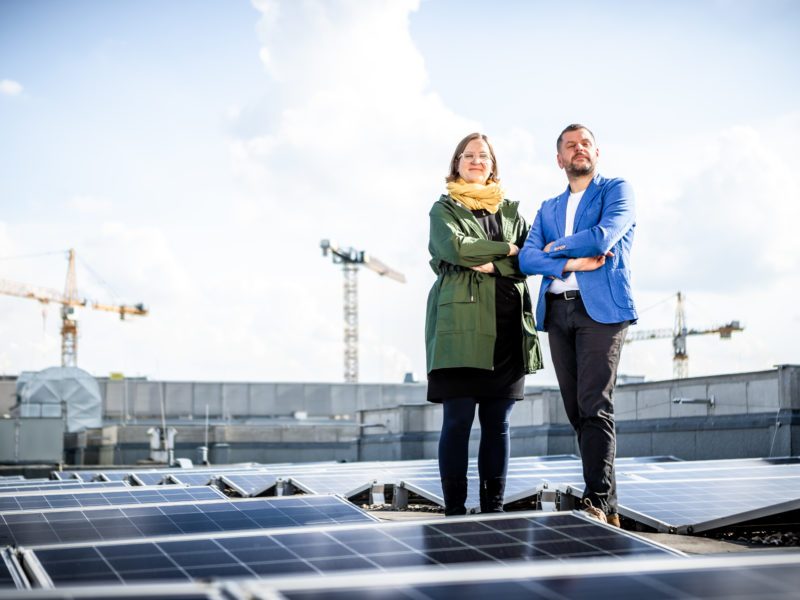 Silke Gebel und Werner Graf stehen auf einem Flachdach, um sie herum Solarpanels. Im Hintergrund der blaue Himmel und ein Kran. Sie haben die Arme verschränkt und lächeln entschlossen.