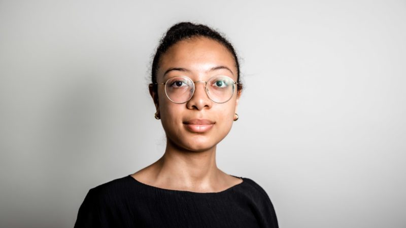 Porträt von Laila Hounkonnou. Sie ist eine junge Frau mit dunklen, zurückgebundenen Haaren. Ihr Blick ist entschlossen in die Kamera. Sie trägt eine runde Brille und ein schwarzes schlichtes Oberteil.