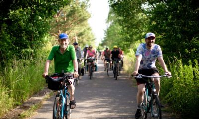 Zwei Männer fahren mit dem Fahrrad frontal auf den Zuschauenden zu. Es ist Sommer, sie tragen kurze Kleidung. Hinter ihnen sind grüne Bäume und unscharf noch viel mehr Radfahrende.