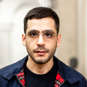 Porträt von Ario Mirzaie. Er schaut entschlossen in die Kamera. Er trägt ein schwarzes T-Shirt und eine dunkelblaue Jacke mit rot kariertem Kragen.