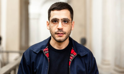 Porträt von Ario Mirzaie. Er schaut entschlossen in die Kamera. Er trägt ein schwarzes T-Shirt und eine dunkelblaue Jacke mit rot kariertem Kragen.