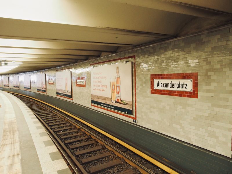 Zu sehen ist ein Ausschnitt des U-Bahnhofes Alexanderplatz von innen. Links ist ein Teil des Bahnsteiges zu sehen. Dann folgt das Bahngleis. Dahinter ist die Wand in grau-weißem Muster. Abwechselnd sind Werbetafeln und ein weißes Schild mit schwarzer Schrift angebracht. Auf dem Schild steht "Alexanderplatz". Es ist umrahmt von roten Backsteinen.