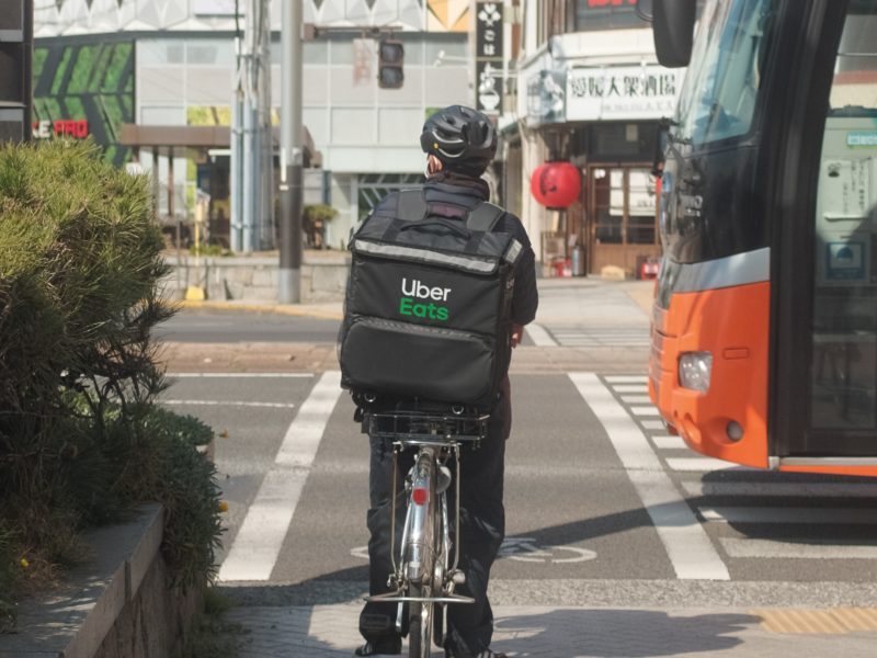 Ein Fahrradfahrer mit einem Rucksack, in dem Essen ausgefliefert wird, fährt auf einem Radstreifen. Von rechts kommt ein Bus angefahren.