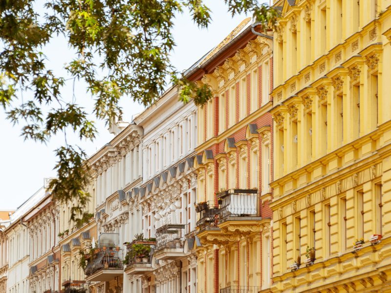 Auf dem Gebäude sind Altbauten schräg von der Seite zu sehen. Eine Fassade ist beige, eine gelb und mehrere weiß. Die Häuser haben Balkone mit vielen Pflanzen. Von oben ragen die Blätter eines Baums ins Bild.
