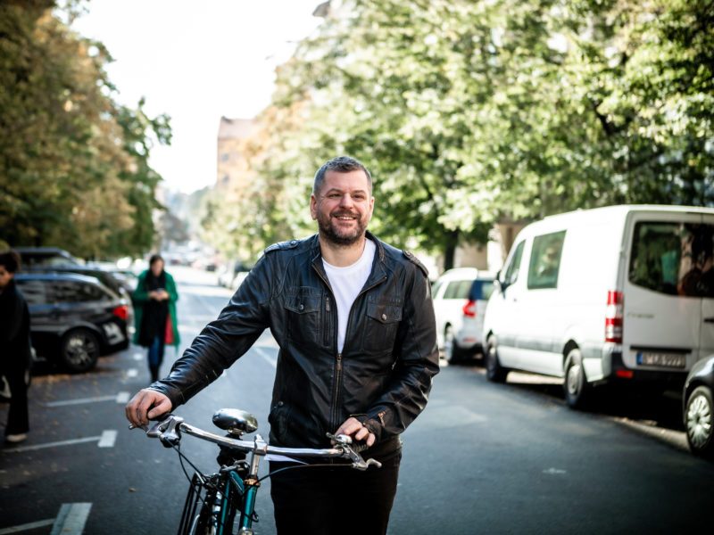 Werner Graf steht in einer Berliner Straße und schiebt ein Fahrrad. Er trägt eine Lederjacke und lächelt in die Kamera.