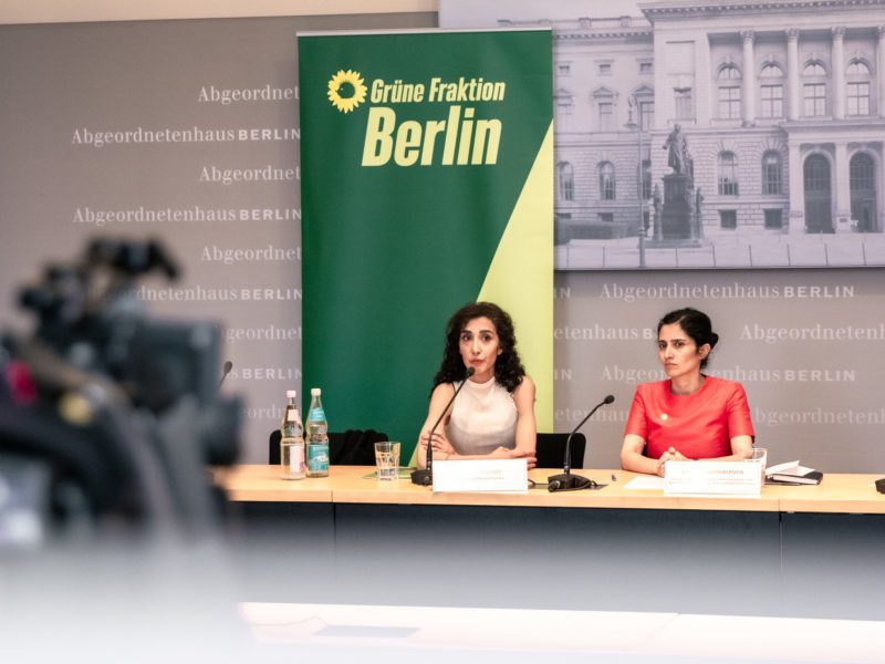 Zwei Frauen sitzen nebeneinander an einem Tisch, sie sind jung. Hinter ihnen steht ein Roll-Up, auf dem "Grüne Fraktion Berlin" steht und im Vordergrund ist eine Kamera zu sehen. Die Szenerie ist eine Pressekonferenz.