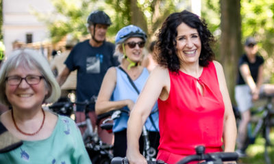 Bettina Jarasch steht mit ihrem Fahrrad in einer Menschenmenge. Sie trägt einen knallroten Jumpsuit und lacht.