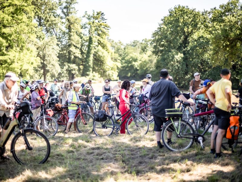 Eine Ansammlung von vielen Fahrradfahrern auf einer Wiese. Es sind bestimmt 30 Personen auf dem Bild, im Hintergrund Bäume.