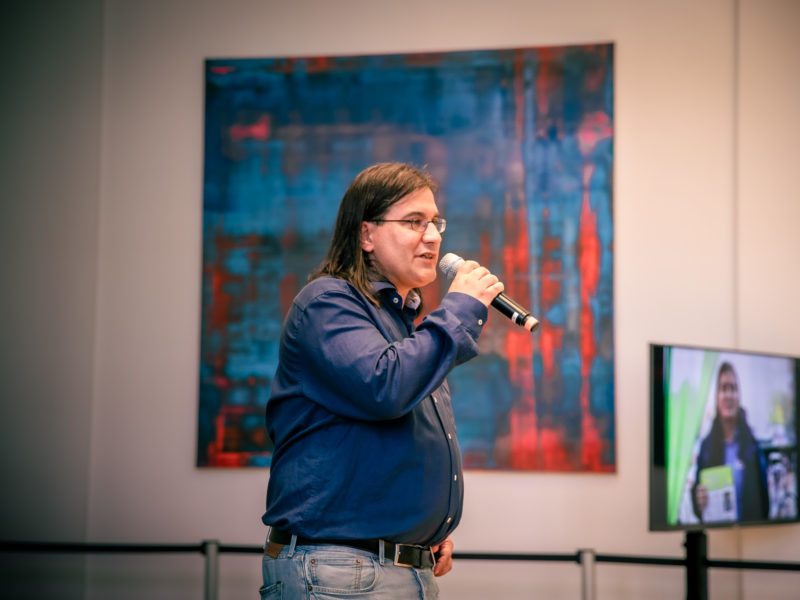 Alexander Kaas-Elias steht vor einem Gemälde und spricht in ein Handmikrofon.