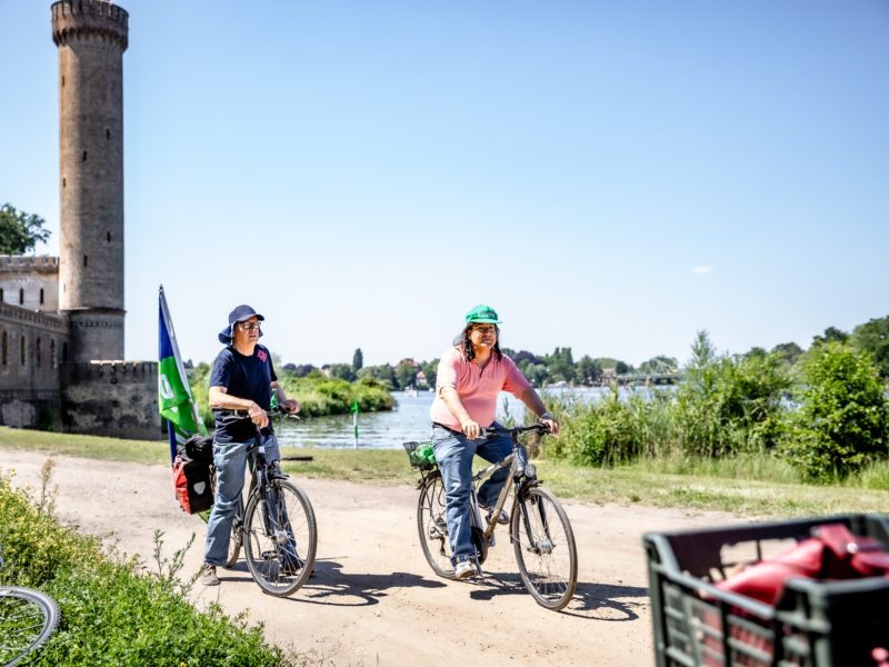Zwei Fahrradfahrer in der Natur. Im Hintergrund ein Fluss und ein alter Backsteinturm.