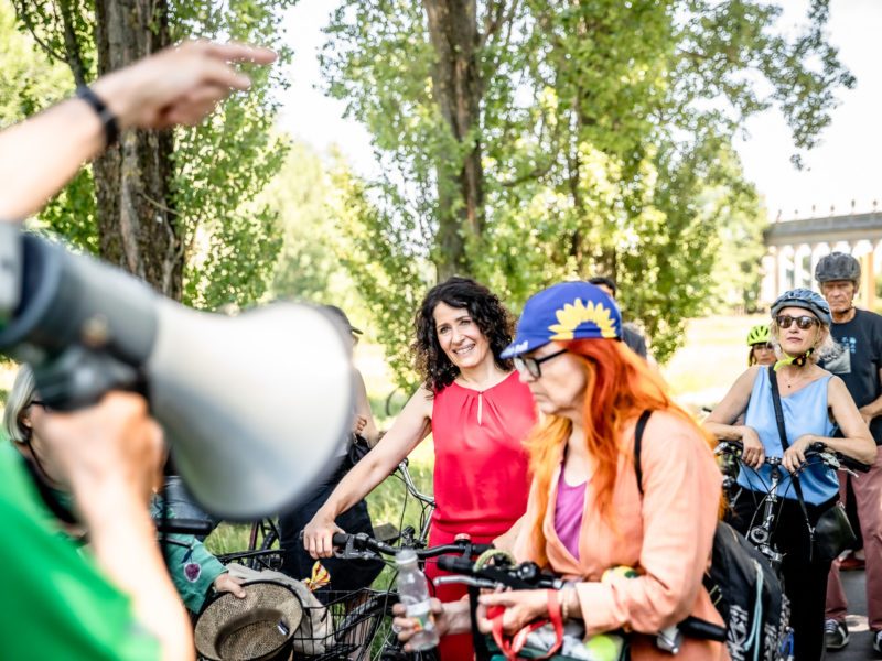 Bettina Jarasch steht mit ihrem Fahrrad in einer Ansammlung von Menschen. Im Vordergrund des Bildes sieht man ein Megafon.