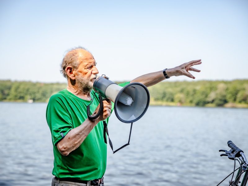 Michael Cramer steht vor einem Fluss und spricht in ein Megafon. Er ist ein 74-jähriger Mann und trägt ein grünes Shirt. Eine Hand hat er gestikulierend ausgestreckt.