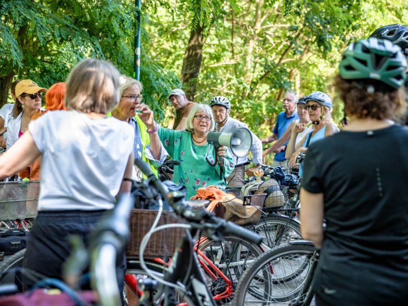 Eine große Gruppe von Menschen mit ihren Fahrrädern steht im Grünen. Inmitten der Gruppe eine ältere Frau, die in ein Megafon spricht.