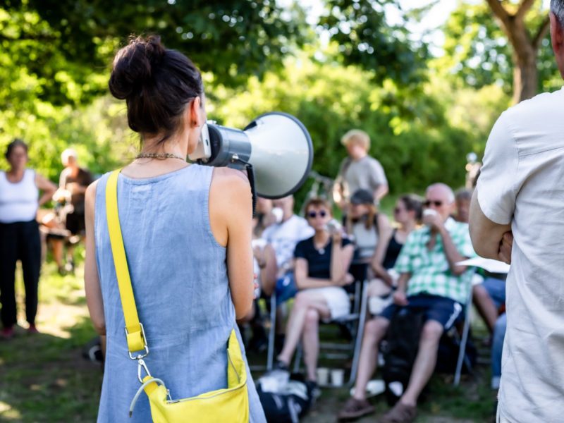 Eine junge Frau mit einem Megafon steht mit dem Rücken zur Kamera und spricht zu einer Gruppe von Menschen, die auf Klappstühlen auf einer Wiese sitzen.