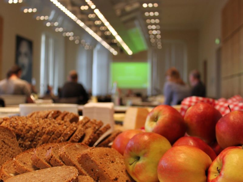 Im Vordergrund des Bildes viele Scheiben aufgeschnittenes Brot und einige Äpfel, im Hintergrund verschwommen ein Raum in dem mehrere Personen sitzen.