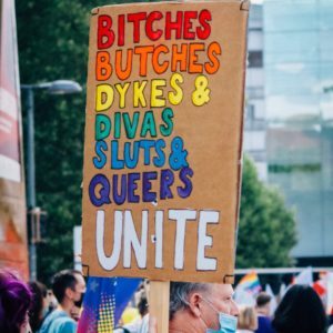 Selbstgebasteltes Pappschild, auf dem in Regenbogenfarben steht: "Bitches, Butches, Dykes & Divas, Sluts & Queers UNITE". Unten im Bild eine Menschenmenge.