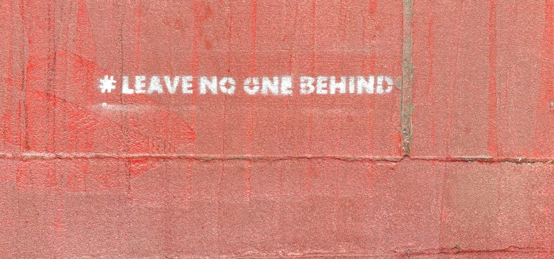 Eine rote Mauer, auf der mit einem Stencil in weiß der Satz "#LEAVE NO ONE BEHIND" aufgesprüht ist.