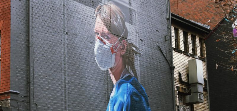 Ein Graffiti an einer Hauswand, das eine Krankenschwester in blauem Kittel und mit Mund-Nasen-Schutz zeigt. Ihr Blick ist angespannt-wütend.