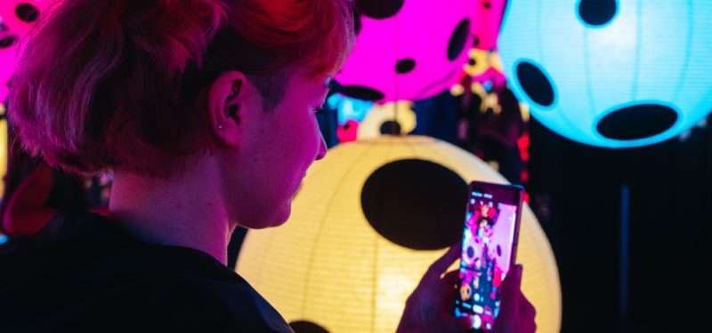 Eine junge Frau steht in einer Kunstausstellung und macht mit ihrem Handy ein Foto. Es sind leuchtende Ballons in verschiedenen Farben mit schwarzen Punkten darauf.