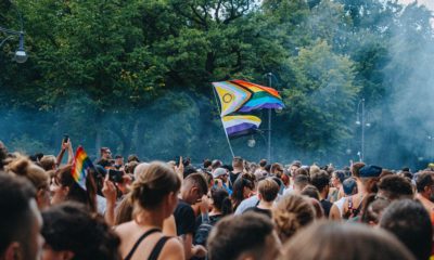 Pride in Berlin. Eine große Ansammlung von Menschen und im Hintergrund Bäume. Im Zentrum des Bildes eine progressive Pride Flag, die hochgehalten wird. Darunter noch eine Non-binary Flag.