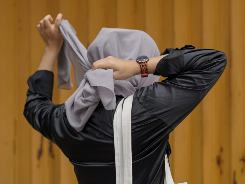 Eine Frau von hinten ist zu sehen, die sich gerade ihren Hijab neu bindet. Sie trägt ein schwarzes Oberteil, eine Tasche über der Schulter und eine Uhr am Handgelenk. Ihr Gesicht ist nicht zu sehen.