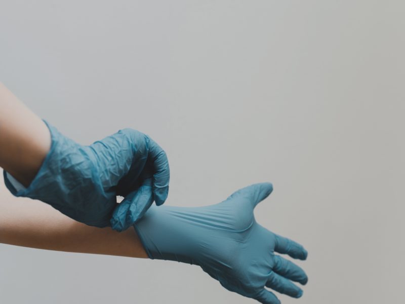 Zwei Hände in blauen Plastikhandschuhen. Eine Hand zieht der anderen gerade den Handschuh an.