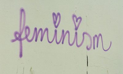 Ein Graffiti-Schriftzug auf einer hellen Wand: "feminism" in lila Schreibschrift. Davor stehen zwei Bistro-Stühle.