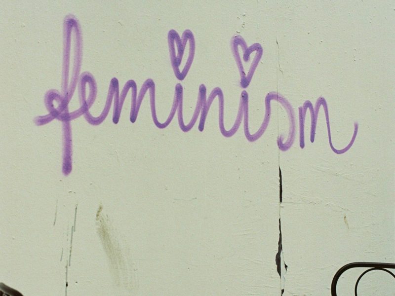 Ein Graffiti-Schriftzug auf einer hellen Wand: "feminism" in lila Schreibschrift. Davor stehen zwei Bistro-Stühle.