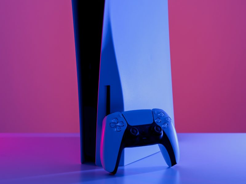 Ein Gaming-Controller vor einer Konsole. Eher Grafik als Foto, in sehr bunten, knalligen Farben (pink und blau).