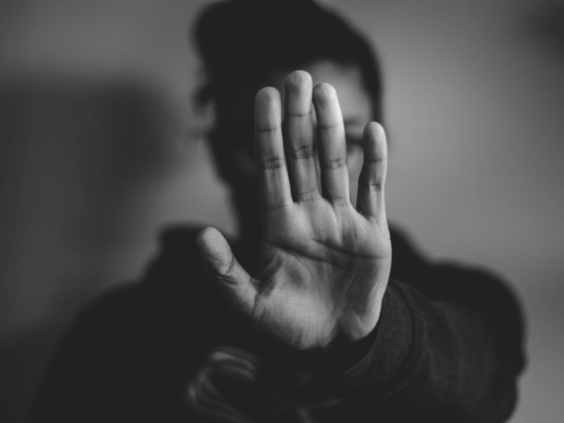 Schwarz-weiß Foto von einer Frau, die ihre Hand zum Schutz vor sich ausstreckt. Ihr Gesicht wird somit durch die Hand verdeckt. Die Handfläche zeigt zum Betrachter.