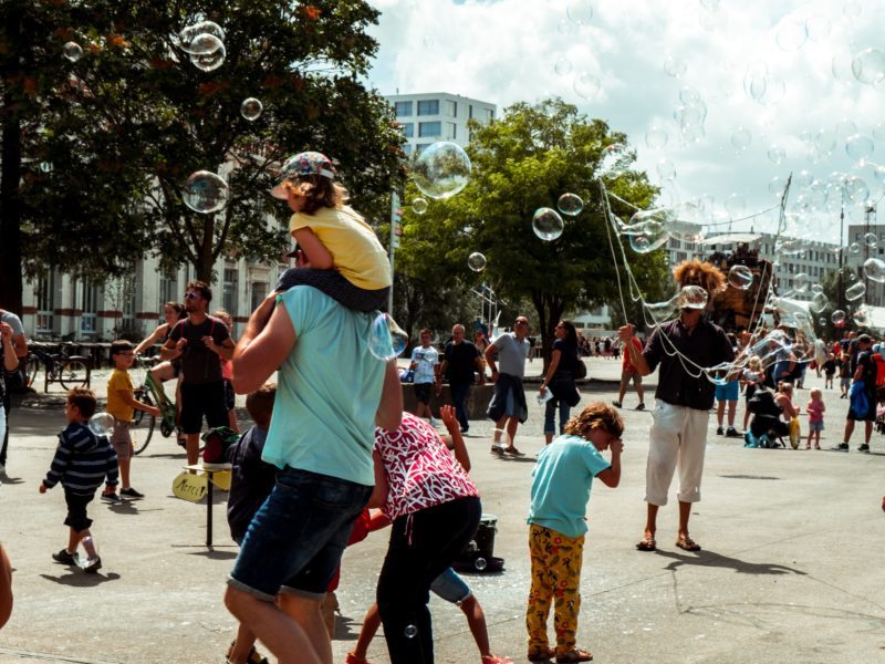 Ein Platz auf dem viel los ist: Viele Menschen, auch viele Kinder laufen herum. Ein Mensch macht große Seifenblasen. Sommerstimmung.