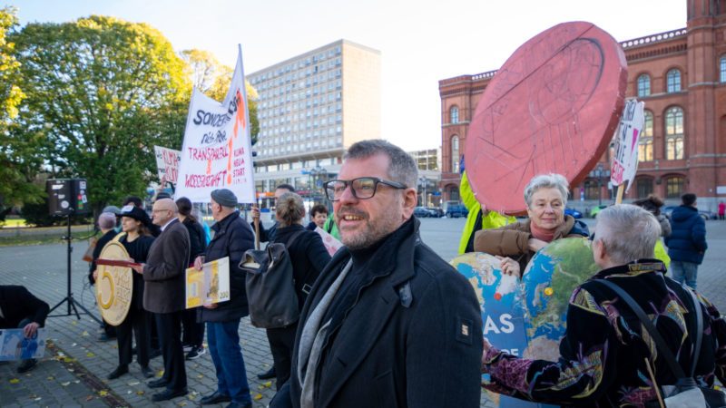 Werner Graf steht in einer Demonstration, er lächelt. Hinter ihm Menschen, die Schilder und Transparente hoch halten